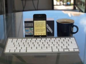 An iPhone showing the text of a note about itself,  with a wireless Bluetooth keyboard and a coffee mug, on a glass table.
